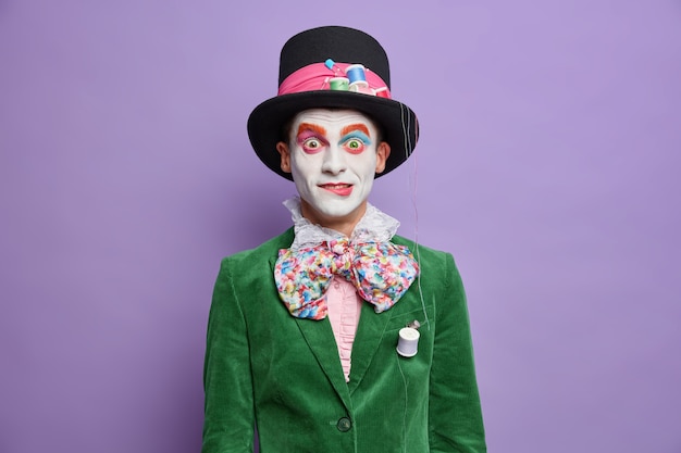 Un participant au festival de parade surpris a l'image d'un chapelier du pays des merveilles porte un maquillage lumineux habillé en costume de carnaval pose contre un mur violet vif