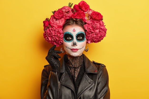 Photo gratuite une participante de vacances mexicaines a un maquillage professionnel, a les yeux noirs porte une couronne de pivoines rouges habillée comme des modèles spirituels à l'intérieur sur un mur vif