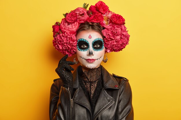 Une participante de vacances mexicaines a un maquillage professionnel, a les yeux noirs porte une couronne de pivoines rouges habillée comme des modèles spirituels à l'intérieur sur un mur vif