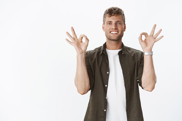 Pas de problème, mon pote, pense que c'est fait, sympathique et joyeux, beau mec blond aux yeux bleus et au sourire parfait montrant un geste correct assurant un plan très apprécié et recommandant le produit sur un mur gris