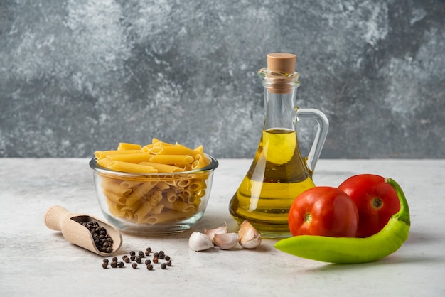 Pâtes crues dans un bol en verre, bouteille d'huile d'olive, grains de poivre et légumes sur tableau blanc.