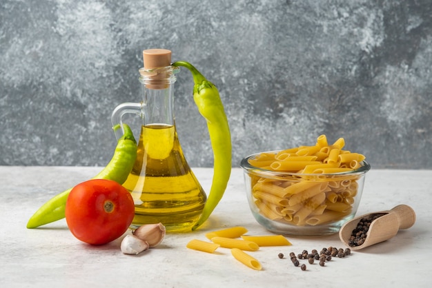 Pâtes crues dans un bol en verre, bouteille d'huile d'olive, grains de poivre et légumes sur tableau blanc.