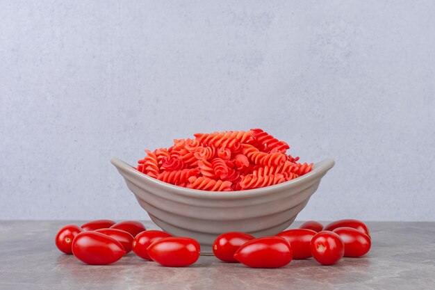 Pâtes fusilli rouges crues dans un bol à côté de tomates, sur la surface en marbre