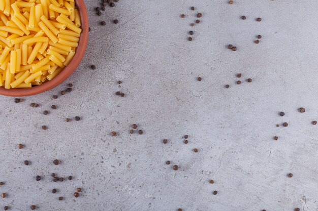 Pâtes italiennes penne séchées dans un bol rond avec des grains de poivre sur une table en pierre.