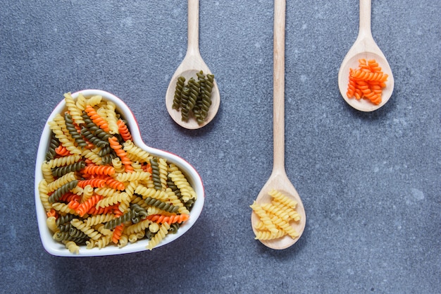 Pâtes macaroni colorées dans un bol en forme de coeur et des cuillères sur une surface texturée grise