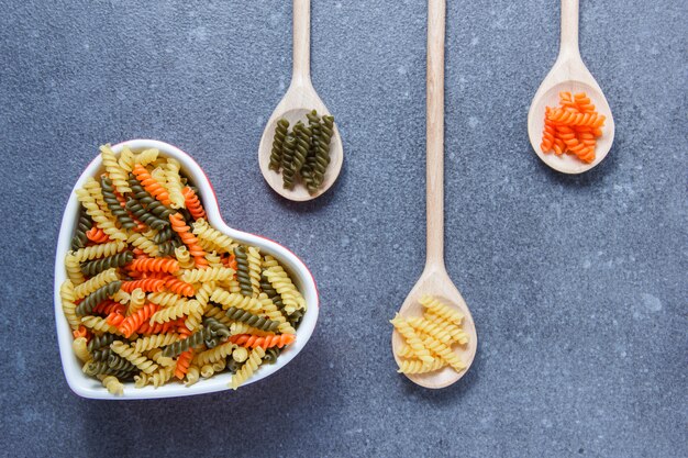 Pâtes macaroni colorées dans un bol en forme de coeur et des cuillères sur une surface texturée grise