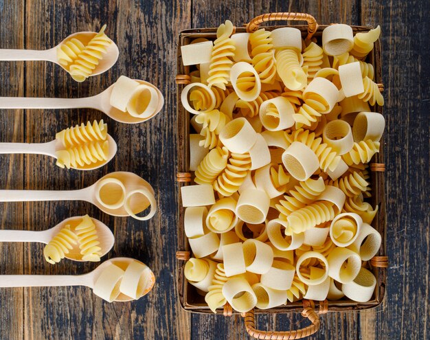 Pâtes macaroni en cuillères et panier sur un fond en bois. vue de dessus.