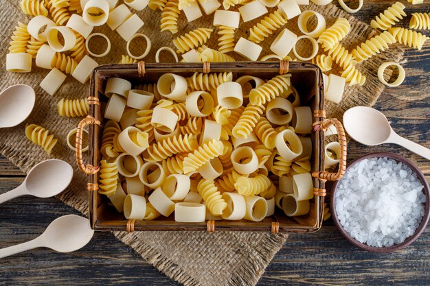 Pâtes macaroni dans un plateau avec des cuillères, vue de dessus de sel sur un sac et fond en bois