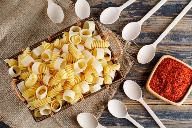 Photo gratuite pâtes macaroni à plat dans un bac avec des cuillères, des épices rouges sur un sac et un fond en bois. horizontal