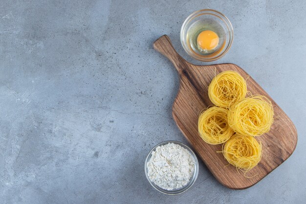 Pâtes nid sèches crues avec oeuf cru et un bol en verre de farine sur un fond de pierre.