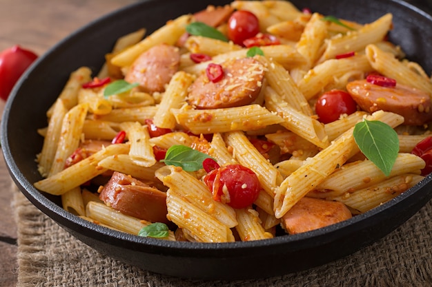 Pâtes penne à la sauce tomate avec saucisse, tomates, basilic vert décorées dans une poêle sur une table en bois