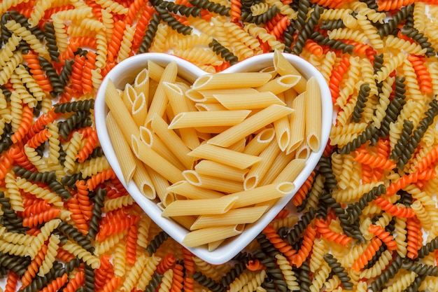 Photo gratuite pâtes penne sèches dans une assiette en forme de coeur à plat sur une table de fusilli de couleur