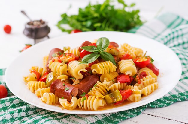 Pâtes à la sauce tomate avec saucisse, tomates, basilic vert décorées dans une assiette blanche sur une table en bois.