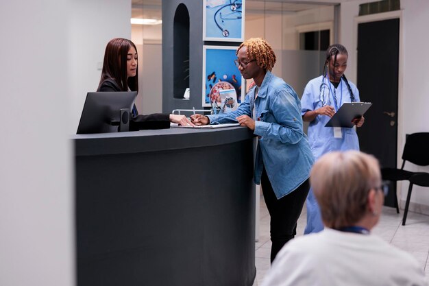 Patient afro-américain remplissant des documents de rapport, parlant à la réceptionniste au comptoir d'accueil de l'hôpital. Femme écrivant un formulaire médical avant un rendez-vous de contrôle avec un médecin au centre de santé.
