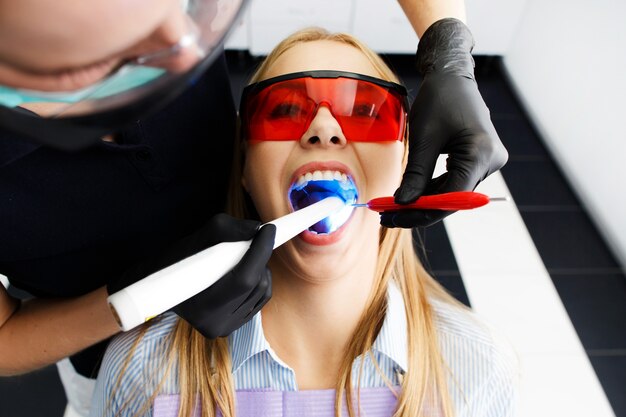 Patient dans des verres rouges est assis sur une chaise au bureau de dentiste pendant que le docteur blanchit ses dents