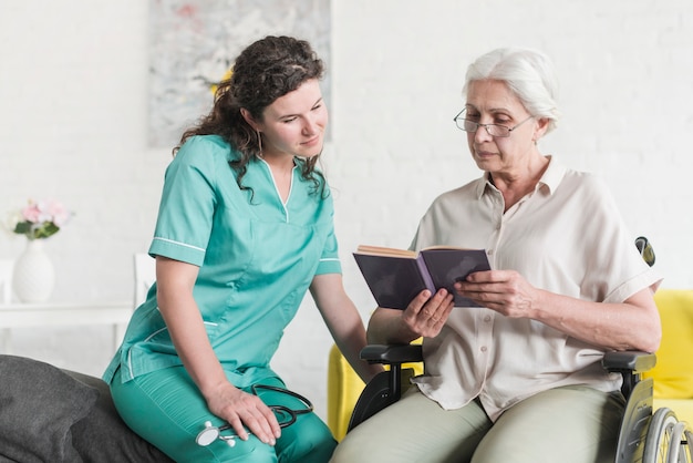 Patient féminin senior handicapé assis sur une chaise roulante, lecture de livre avec infirmière