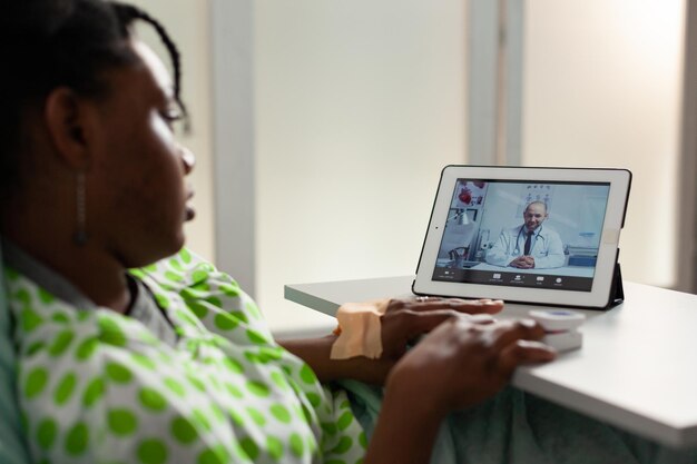 Patiente afro-américaine malade allongée dans son lit discutant d'un traitement médicamenteux avec un médecin à distance lors d'une conférence de réunion par vidéoconférence en ligne. Appel de télésanté sur écran de tablette