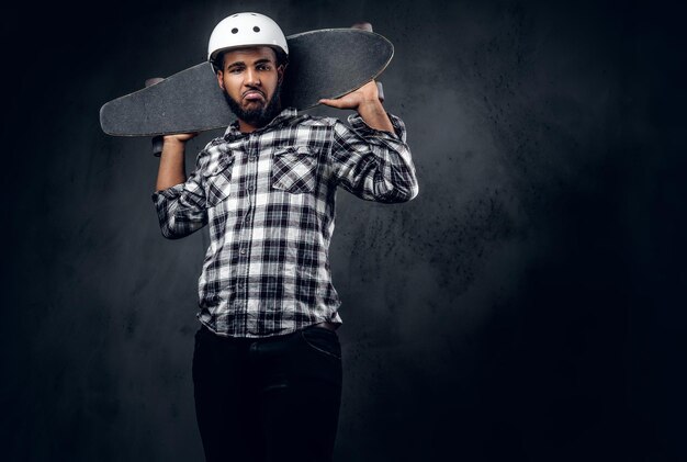 Un patineur noir vêtu d'une chemise polaire tient une longue planche sur fond gris dans un studio.