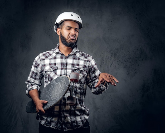 Un patineur noir vêtu d'une chemise polaire tient une longue planche sur fond gris dans un studio.