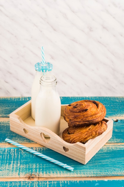 Photo gratuite pâtisseries danoises avec une bouteille de lait sur un plateau en bois près de la paille sur une table en bois bleue