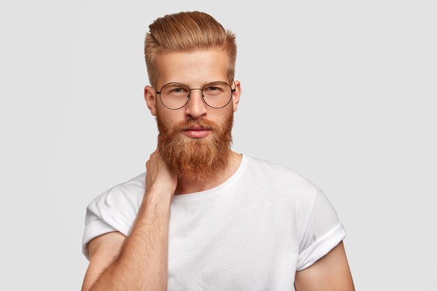 Le patron de bel homme sérieux a une coupe de cheveux à la mode et une barbe de gingembre, garde les mains derrière le cou, regarde avec confiance