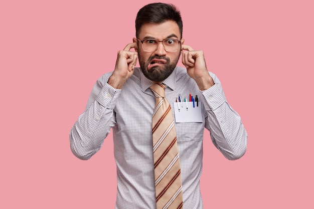 Photo gratuite le patron masculin mécontent et ennuyé est habillé formellement, se bouche les oreilles, ne veut pas entendre les plaintes de ses collègues, fronce les sourcils avec mécontentement