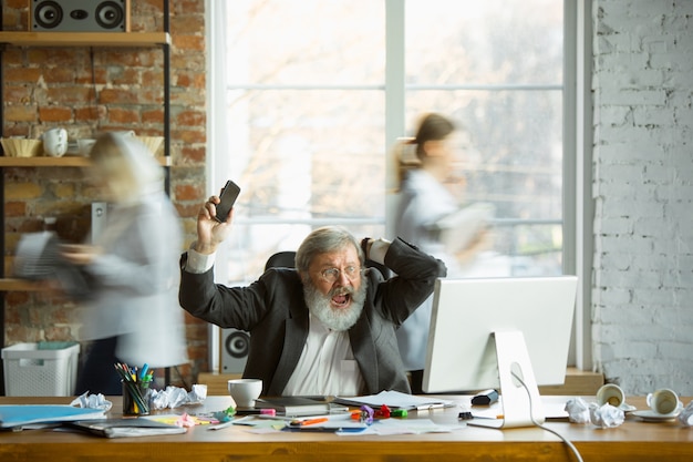 Photo gratuite patron nerveux et fatigué sur son lieu de travail occupé pendant que les gens se déplacent presque flous. employé de bureau, gestionnaire de travail, a des problèmes et des délais, ses collègues distrayants. entreprise, travail, concept de charge de travail.