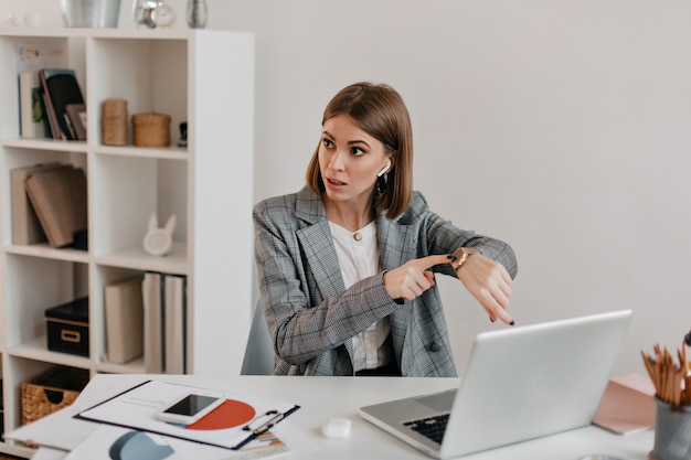 La patronne en tenue élégante fait remarquer à l'horloge, parlant de la fin de son subordonné. Portrait de femme assise dans un bureau léger.