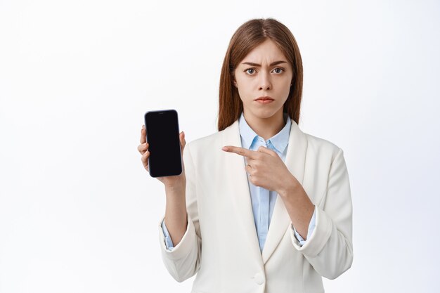 Une PDG de bureau sérieuse et mécontente pointe un écran de smartphone vide, fronçant les sourcils et l'air déçu, debout sur un mur blanc