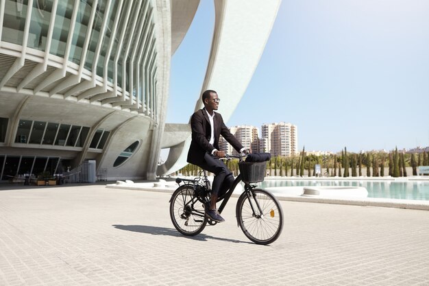 PDG confiant et respectueux de l'environnement à la peau sombre utilisant un véhicule d'assistance à pédale à deux roues pour se rendre au travail. Homme d'affaires noir moderne prospère à vélo au bureau après le déjeuner. Les gens, les transports et les affaires