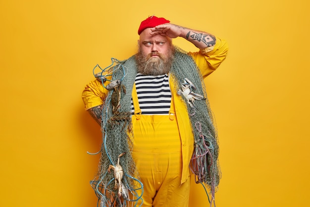 Un pêcheur sérieux barbu garde la main sur le front et regarde au loin, pose avec un filet de pêche sur les épaules, attrape des créatures marines, a un gros ventre, des bras tatoués