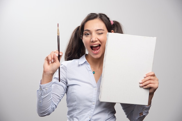 Peintre brune posant avec toile et pinceau sur mur gris.