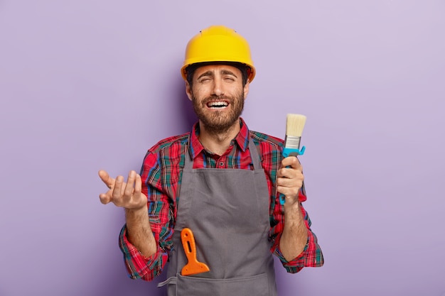 Photo gratuite un peintre masculin mécontent et malheureux tient un pinceau, effrayé d'avoir beaucoup de travail, occupé à réparer à la maison, porte un casque jaune, un tablier gris. ouvrier du bâtiment pose avec des outils de construction