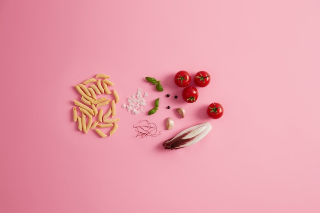 Penne riz sec pâtes basilic, salade de chicorée, tomates, fils de piment rouge à l'ail pour préparer une délicieuse cuisine italienne gastronomique. Macaroni non cuit et ingrédients sur fond rose. La nourriture saine