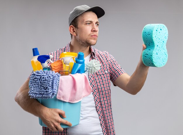 pensant que jeune homme nettoyeur portant une casquette tenant un seau avec des outils de nettoyage et regardant une éponge de nettoyage dans sa main isolée sur un mur blanc