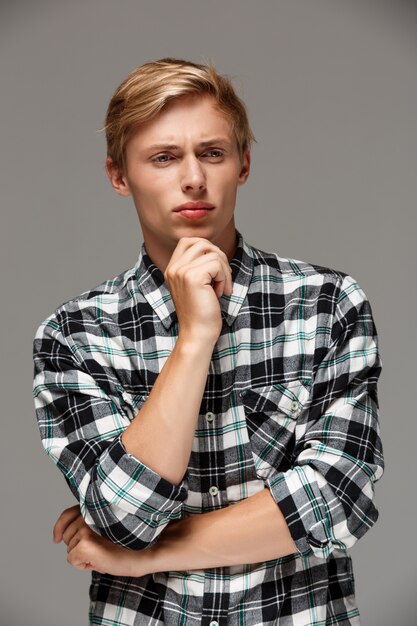 Pensée confiante beau beau jeune homme portant une chemise à carreaux décontractée avec les mains croisées sur la poitrine à la recherche de mur gris