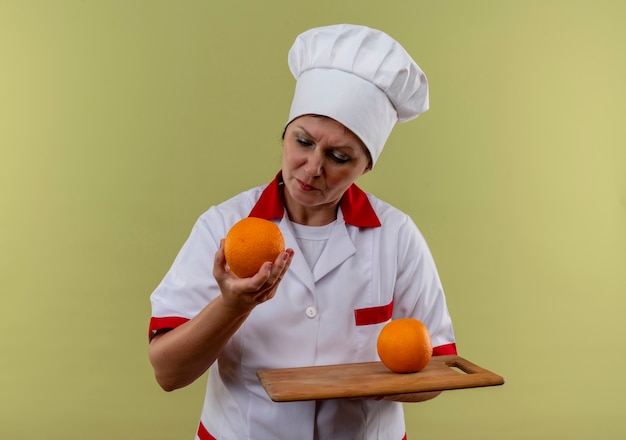 Pensée femme d'âge moyen cuisinier en uniforme de chef tenant orange sur une planche à découper et regardant orange dans sa main