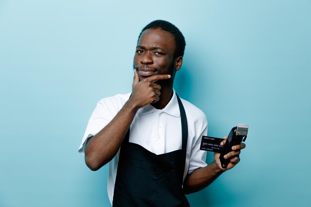 Penser attrapé le menton tenant la carte avec des tondeuses à cheveux jeune coiffeur afro-américain en uniforme isolé sur fond bleu