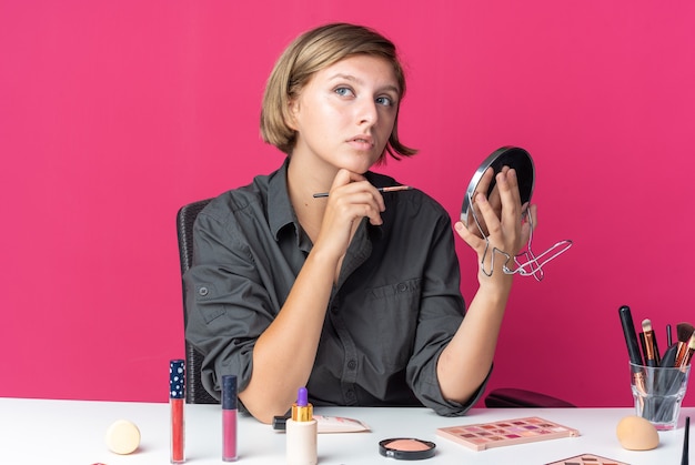Penser une belle jeune femme assise à table avec des outils de maquillage tenant un pinceau de maquillage avec miroir