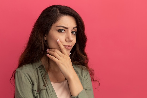 Penser une belle jeune fille portant un t-shirt vert olive saisit le menton isolé sur un mur rose avec espace pour copie