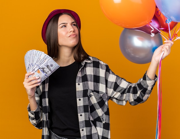 Penser à côté de la belle jeune fille portant un chapeau de fête tenant des ballons avec de l'argent isolé sur un mur orange