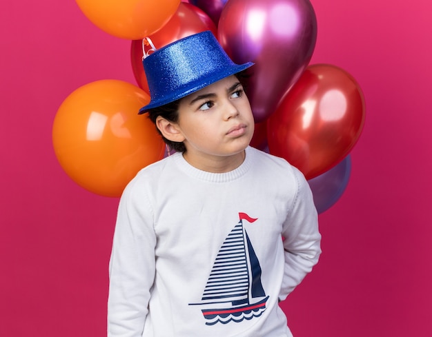 Penser à côté petit garçon portant un chapeau de fête bleu debout devant des ballons isolés sur un mur rose