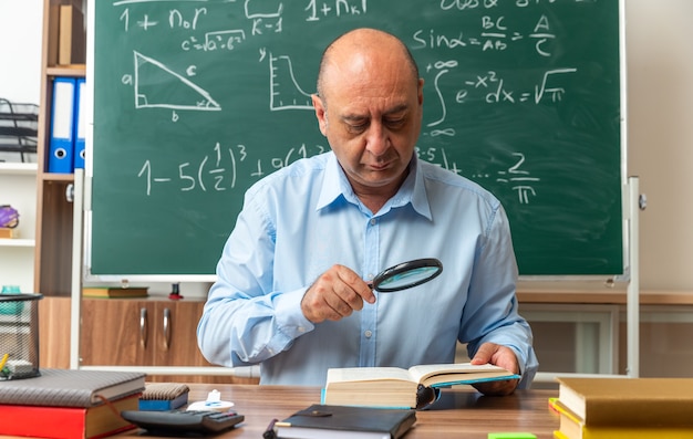 Penser un enseignant masculin d'âge moyen est assis à table avec des fournitures scolaires, un livre de lecture avec une loupe en classe