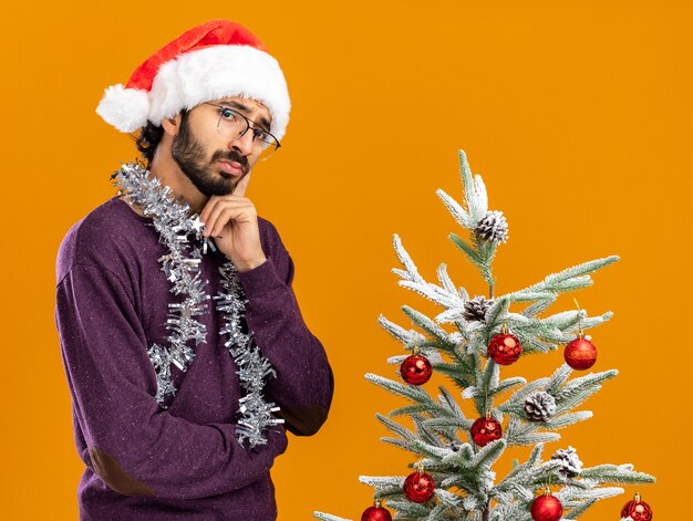 Penser un jeune beau mec debout à proximité d'un arbre de Noël portant un chapeau de Noël avec une guirlande sur le cou mettant le doigt sur la joue isolé sur un mur orange