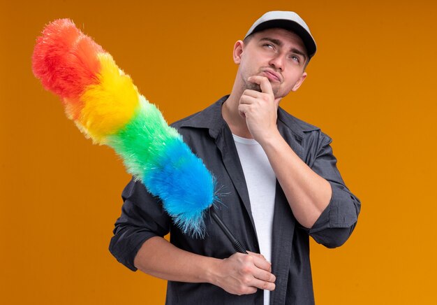 Penser jeune beau mec de nettoyage portant un t-shirt et une casquette tenant pipidaster a attrapé le menton isolé sur le mur orange