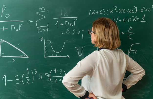 Photo gratuite penser la jeune enseignante debout devant le tableau noir mettant les mains sur la hanche en classe