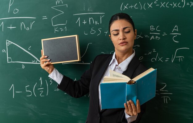 Penser une jeune enseignante debout devant un tableau noir tenant un mini tableau regardant un livre dans sa main en classe