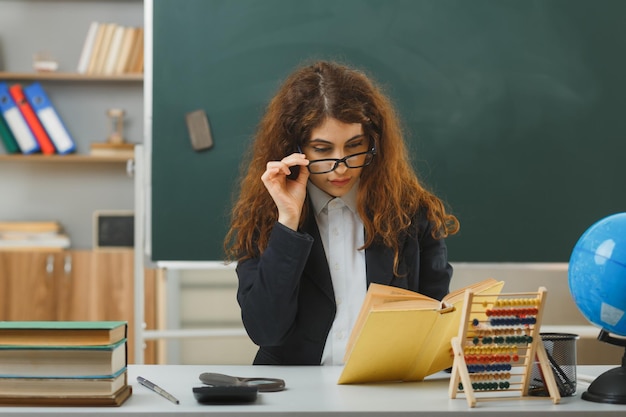 Photo gratuite penser jeune enseignante portant des lunettes tenant et lisant un livre assis au bureau avec des outils scolaires en classe