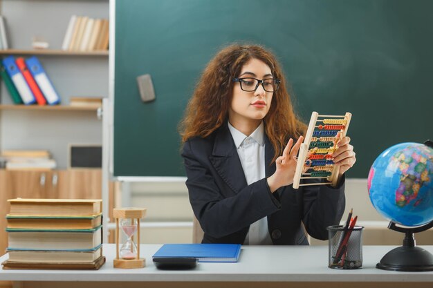 penser jeune enseignante portant des lunettes tenant et regardant l'abaque assis au bureau avec des outils scolaires en classe