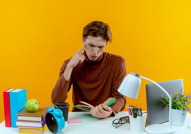 Penser jeune étudiant garçon assis au bureau avec des outils scolaires tenant et regardant livre mettant la main sur le menton sur jaune